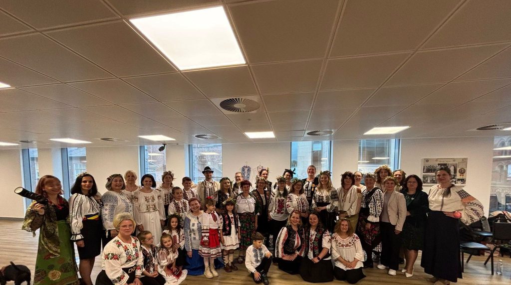 Group photograph from the Universal Day of the Romanian and Moldovan Blouse celebration in Manchester. Members of the Romanian and Moldovan communities, including adults and children, are gathered indoors wearing traditional embroidered blouses known as ii and authentic folk costumes from various regions of Romania and the Republic of Moldova. Participants proudly display their cultural heritage through colourful embroidery, traditional patterns and floral wreaths. The image captures community spirit, cultural diplomacy, intergenerational participation and the preservation of Romanian and Moldovan traditions in the United Kingdom. The event celebrates identity, heritage, unity and the UNESCO-recognised art of the traditional blouse with altiță while strengthening cultural ties between the Romanian, Moldovan and wider local communities in Manchester.